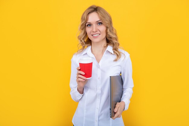 Cheerful blonde woman with coffee cup and laptop on yellow background
