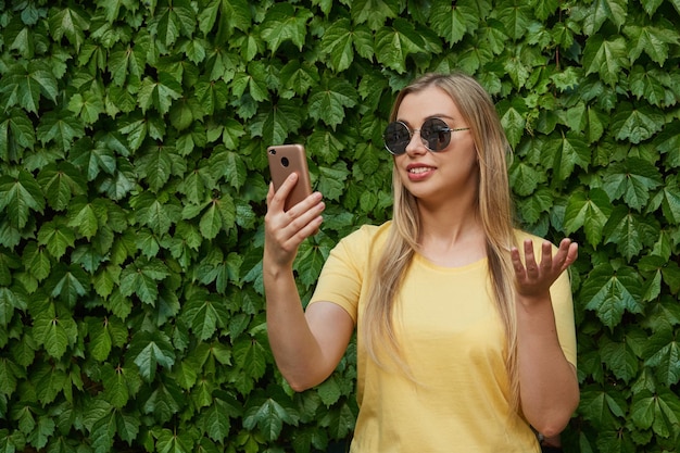 サングラスの黄色いTシャツを着た陽気なブロンドの女性は携帯電話を使用しています