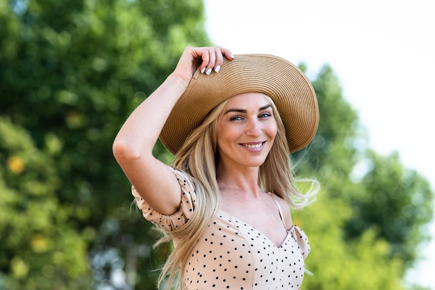 Cheerful blonde young Scandinavian woman in beige dress and straw hat toothy smiling looking at camera against green trees on background Playful Itali girl traveling on vacations having fun Leisure