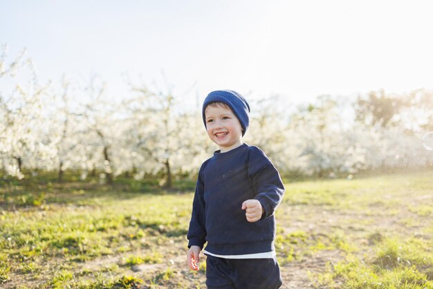 Cheerful boy 3 years old on a walk in a blooming garden a little boy 3 years old in a sweater and a hat runs through a blooming garden clothes for children aged 3 years emotions of child's joy