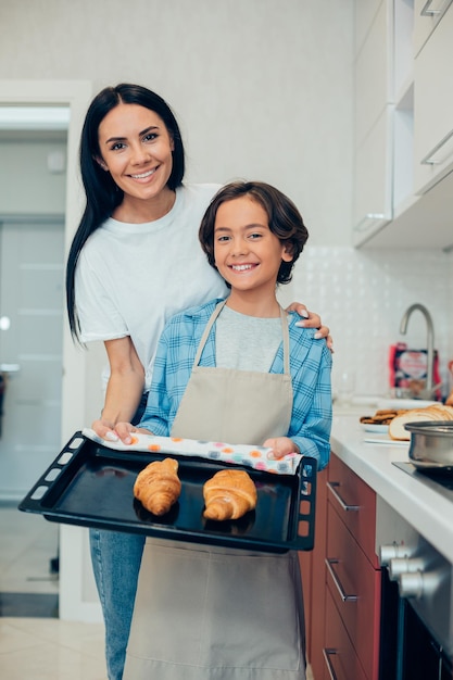 Cheerful boy in apron holding a hot baking pan with delicious croissants while standing in the kitchen with his gladsome mother