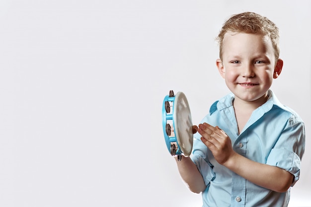 A cheerful boy in a blue shirt holding a tambourine and smiling on a light background