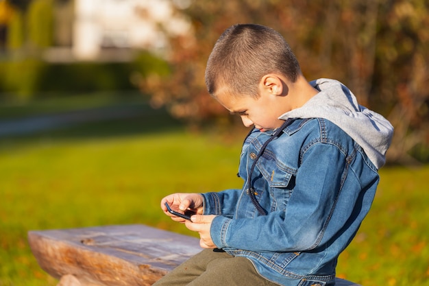 A cheerful boy in a denim jacket lies on a bench and plays with a smartphone in a city park