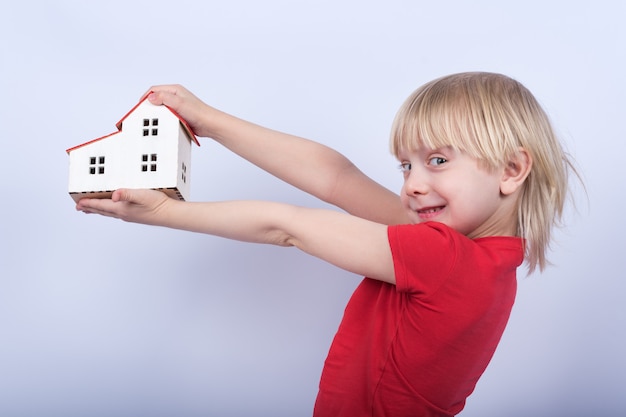 Cheerful boy holding model house and laughs. portrait of child
with toy house in hands on white background.