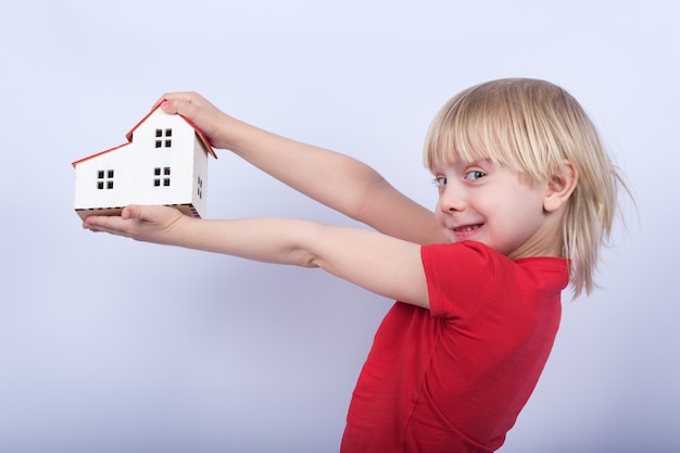 その陽気な男の子はモデルの家と笑いを保持しています。白い背景の上の手の中のおもちゃの家を持つ子供の肖像画。