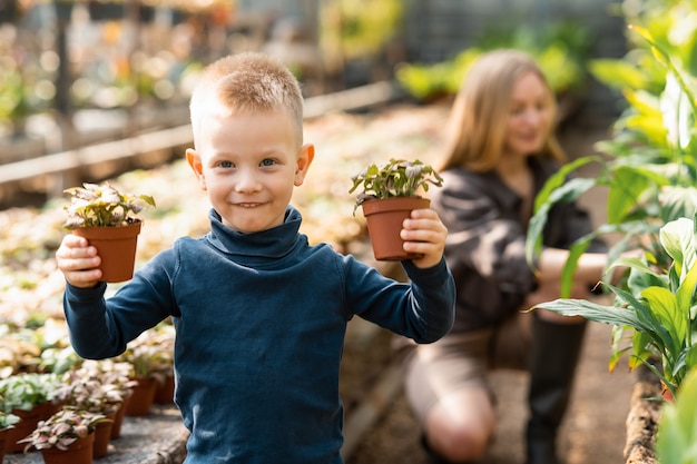 Веселый мальчик держит в руке горшки с растениями во время прогулки в теплице с мамой