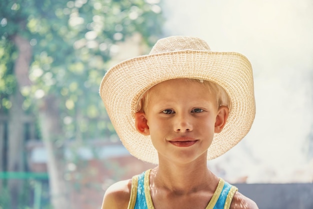 Cheerful boy in large straw hat and blue sleeveless shirt posing for camera in summer city park