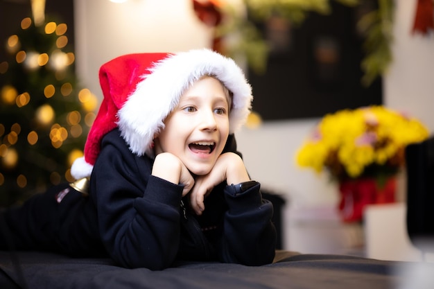 A cheerful boy laughs in a Santa Claus hat wrung his hands at his chin and looks away