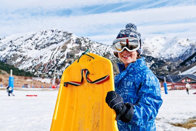 Cheerful boy in outerwear with yellow plastic sleigh smiling and looking at camera while standing against snowy mountain ridge on sunny day on ski resort