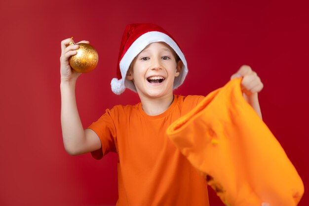 Un ragazzo allegro con un cappello da babbo natale e un sacchetto regalo di natale ha tirato fuori un giocattolo per la palla dell'albero di natale