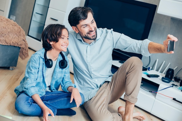 Cheerful boy sitting with his father on the floor and smiling
positive man taking selfies