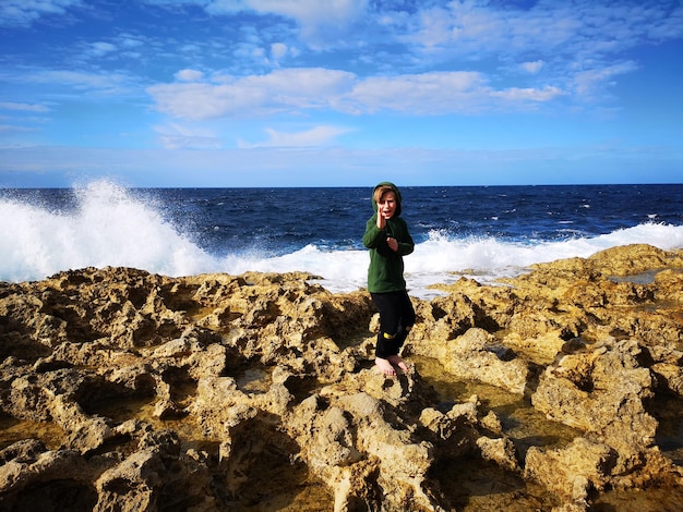 Cheerful boy standing on rock at beach against sky