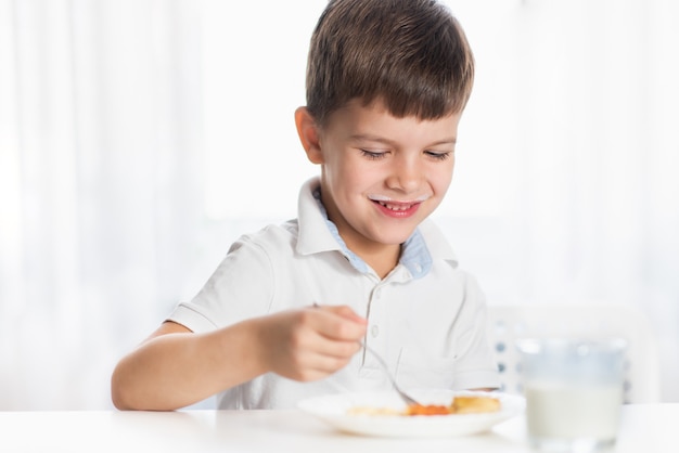 Cheerful boy in white shirt eats cottage cheese pie and drinks milk for breakfast at home