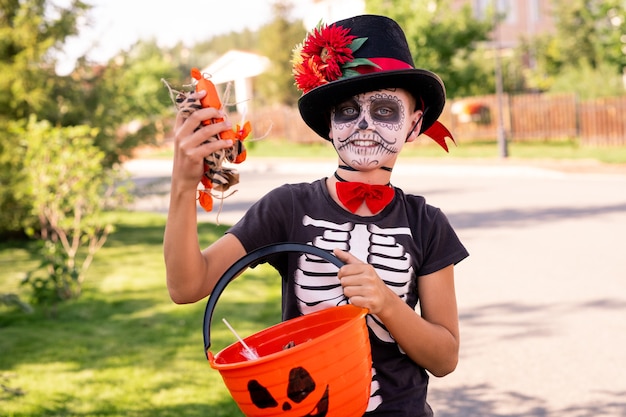 Cheerful boy with painted face in halloween t-shirt and elegant hat holding plastic basket and boastin with sweet treats in front of camera