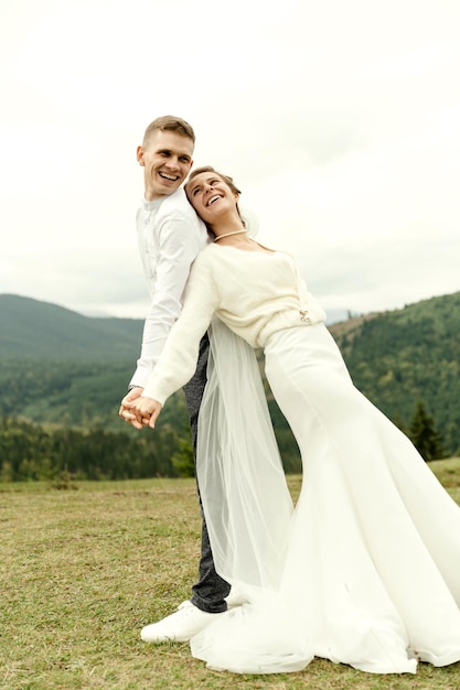 Cheerful bride and groom stand with their backs to each other and hold hands on the top of the mountain wedding day of a young couple in love