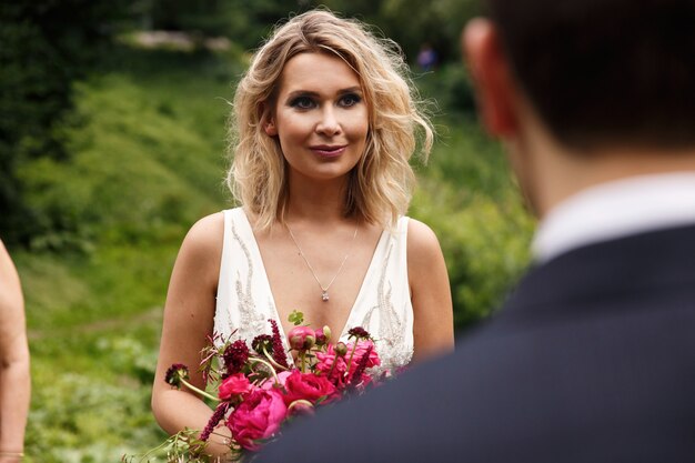 Cheerful bride stands with red wedding bouquet before a groom