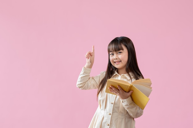 A cheerful and bright young Asian girl is holding an opened book and pointing her finger up