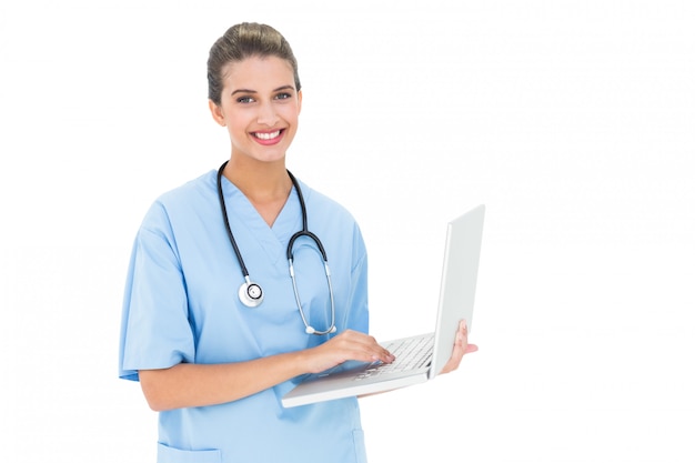 cheerful brown haired nurse in blue scrubs using a laptop