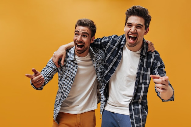 Cheerful brunet happy men in white tshirts and blue checkered shirts point at camera and laugh on isolated orange background