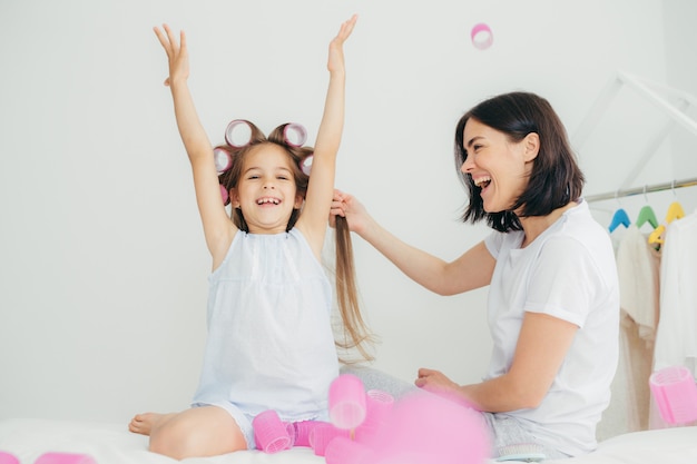 Cheerful brunette female does hair of her daughter who has glad expression, raised hands and has curlers on head