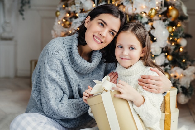 Cheerful brunette female leans at her daugter, embraces her, presents gift box, being in living room near decorated New Year tree. Glad family: mother and daughter in warm sweaters celebrate Christmas