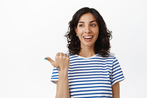 Cheerful brunette girl laughing, smiling white teeth and pointing left, looking at logo banner with satisfied face, recommend company, standing over white background