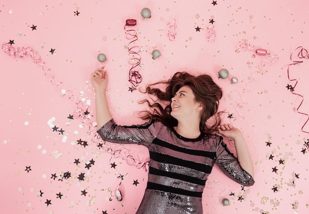 Cheerful brunette girl lies on a pink background among confetti and streamer, the concept of celebrating christmas and new year, festive party.