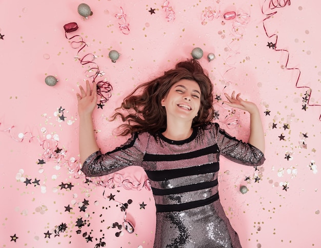 Cheerful brunette girl lies on a pink background among confetti and streamer, the concept of celebrating christmas and new year, festive party.