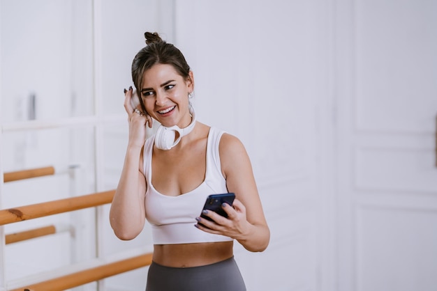 Photo cheerful brunette girl in sportswear holds phone smiles checking sound of new headphones at fitness