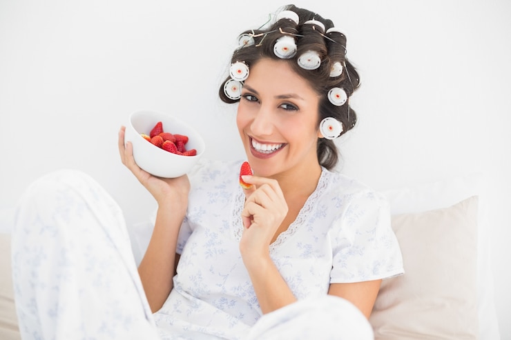 Photo cheerful brunette in hair rollers having a bowl of strawberries