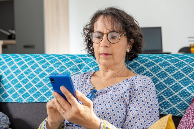 Donna senior castana allegra che per mezzo dello smartphone mentre sedendosi sul sofà