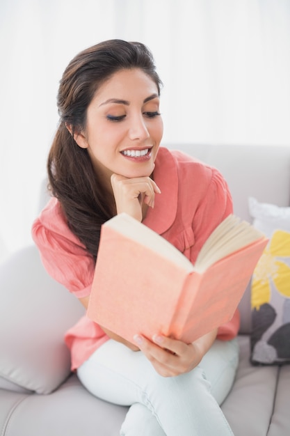 Brunette allegro che si siede sul suo sofà che legge un libro