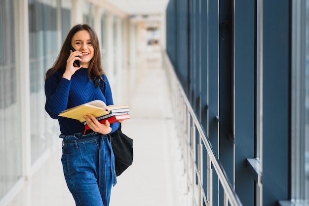 黒のバックパックと陽気なブルネットの学生の女の子は、大学の廊下で本を立っている近代的な建物の女子学生で本を保持します