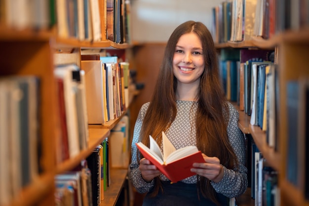Веселая брюнетка-студентка, стоящая с книгой в руках между рядами книжных полок в библиотеке, читающая для удовольствия, получающая знания и наслаждающаяся образованием