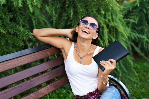 Cheerful brunette woman in casual clothes and sunglasses sits on a wooden bench in the park against ...