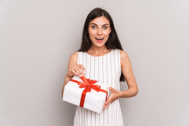  cheerful brunette woman wearing dress smiling and holding present box isolated over gray wall