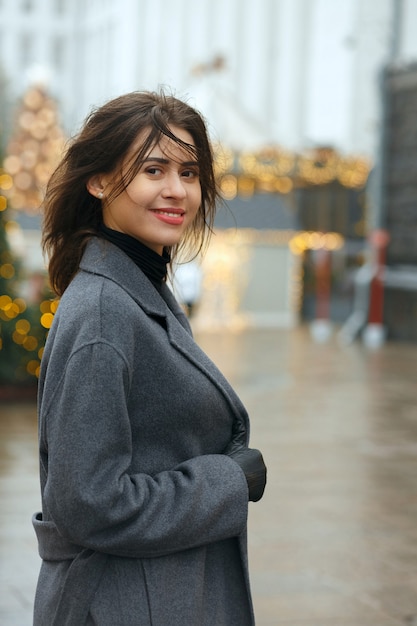 Cheerful brunette woman wears trendy coat walking in a rainy day at the city decorated to Christmas