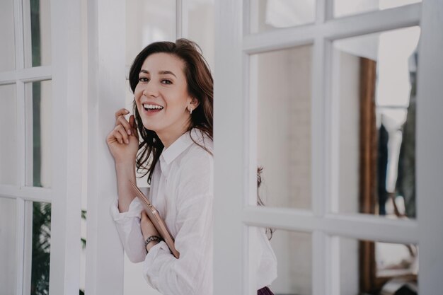 Cheerful brunette woman in white shirt carrying laptop toothy smiling looks happily completed work