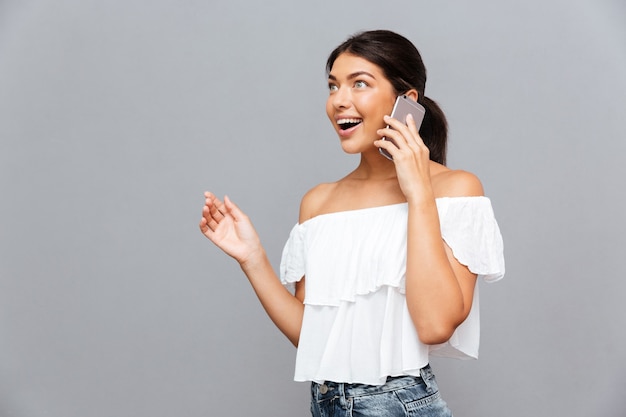 Cheerful brunette young woman talking on the phone isolated on a gray wall