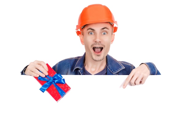 A cheerful builder in a denim jacket and with an orange helmet on his head is holding a gift in his hand on an isolated white background. Emotional photo. Copy space.