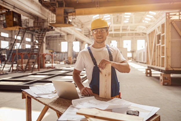 Cheerful builder standing by the table with laptop and papers