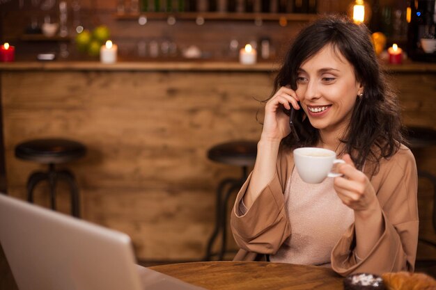 陽気なビジネスウーマンは微笑んで、コーヒーショップで彼女のラップトップを見ています。コーヒーショップでポータブルコンピューターを持っている女の子。スマートフォンで話している若いフリーランサー。