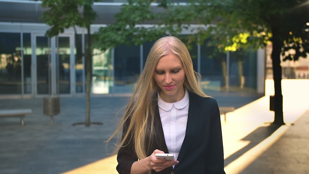 Donna allegra di affari con il telefono all'aperto. lass biondo contento in smartphone della tenuta del rivestimento nero e distogliere lo sguardo con il sorriso sulla via