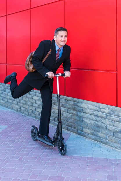 Cheerful businessman quickly rides a kick scooter by the red wall