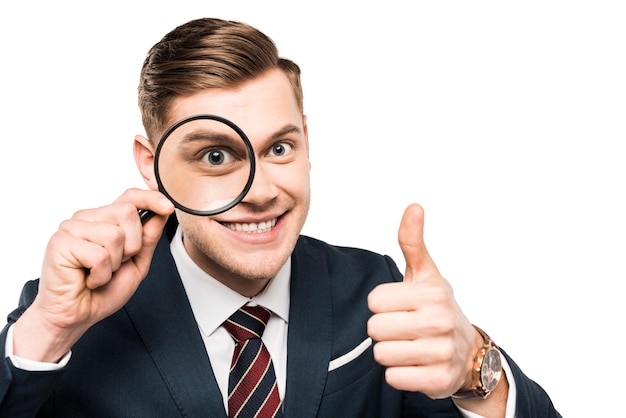 Cheerful businessman showing thumb up and holding magnifying glass near eye isolated on white