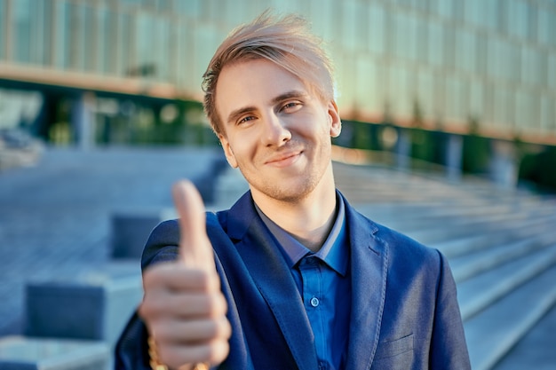 Cheerful businessman in suit shows thumb near stairs of office center