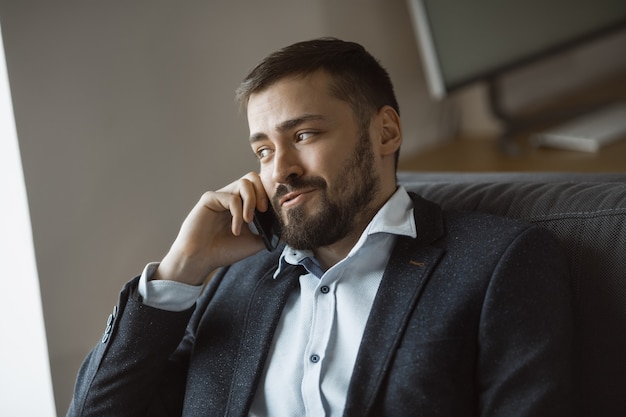 Cheerful businessman in suit talking on the phone, sitting on sofa in office.