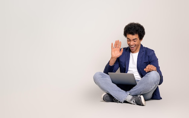 Веселый бизнесмен махает рукой во время онлайн-встречи на ноутбуке, сидя на белом фоне