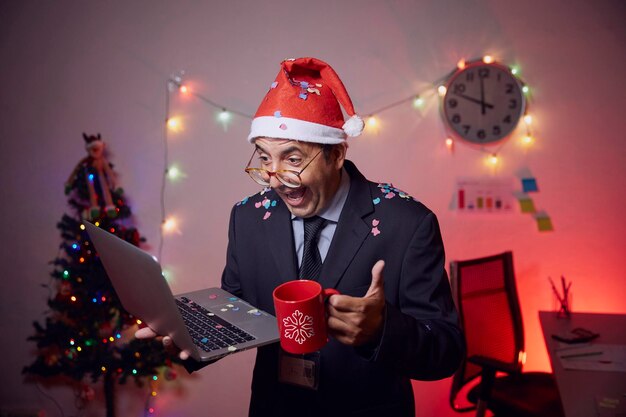 Photo cheerful businessman wearing santa hat holding laptop and coffee cup against illuminated christmas tree and lights at home