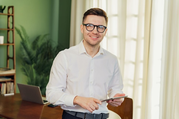 Cheerful businessman in a white shirt using a tablet in a sunny room with a laptop on the desk